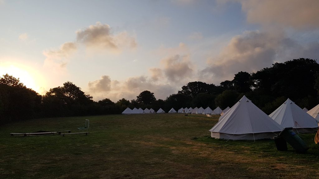 White bell tents in a field surrounded by trees. Cloudy sky with sun rising.